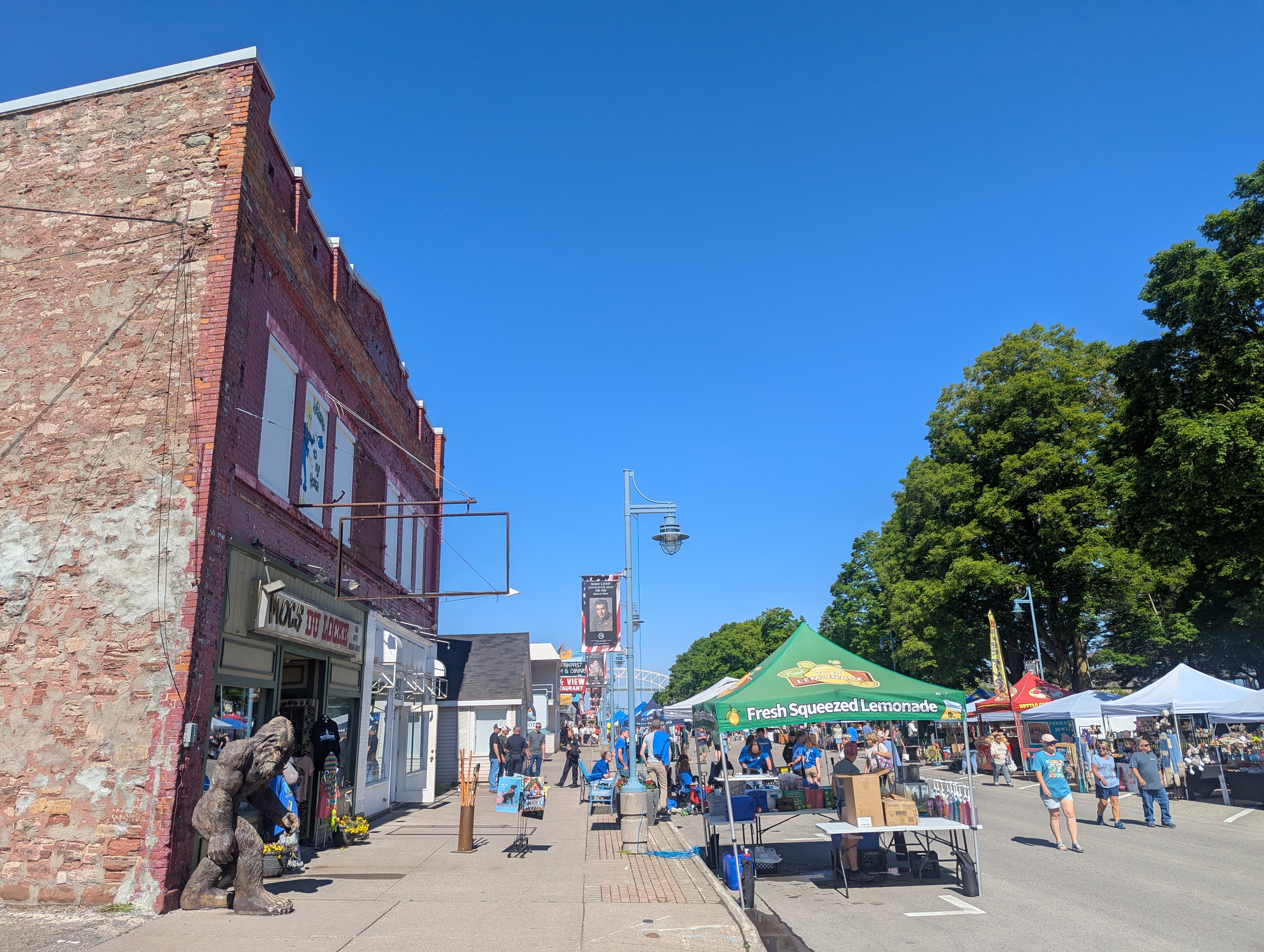 Engineers Day celebrations in downtown Sault Ste. Marie. Buildings are visible on the left side of the street, while tents are set up on the street, with the one near the front offering "Fresh Squeezed Lemonade". There are a number of people walking around, but it is not crowded.