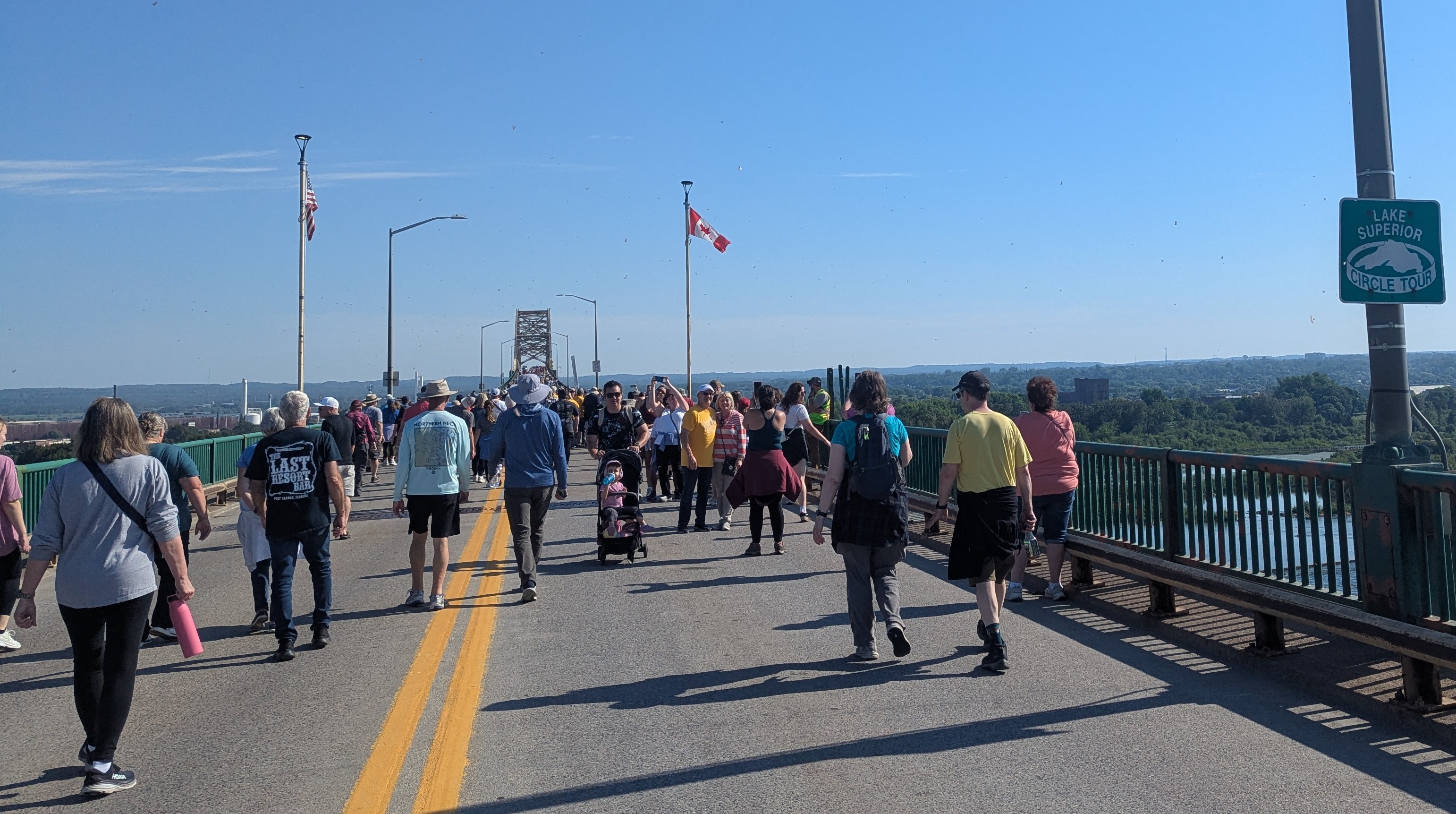 A view from the bridge walk. Both the American and Canadian flags are visible ahead, denoting the international border. A reassurance shield for the Lake Superior Circle Tour is visible on the right. A significant number of people are walking on the bridge.